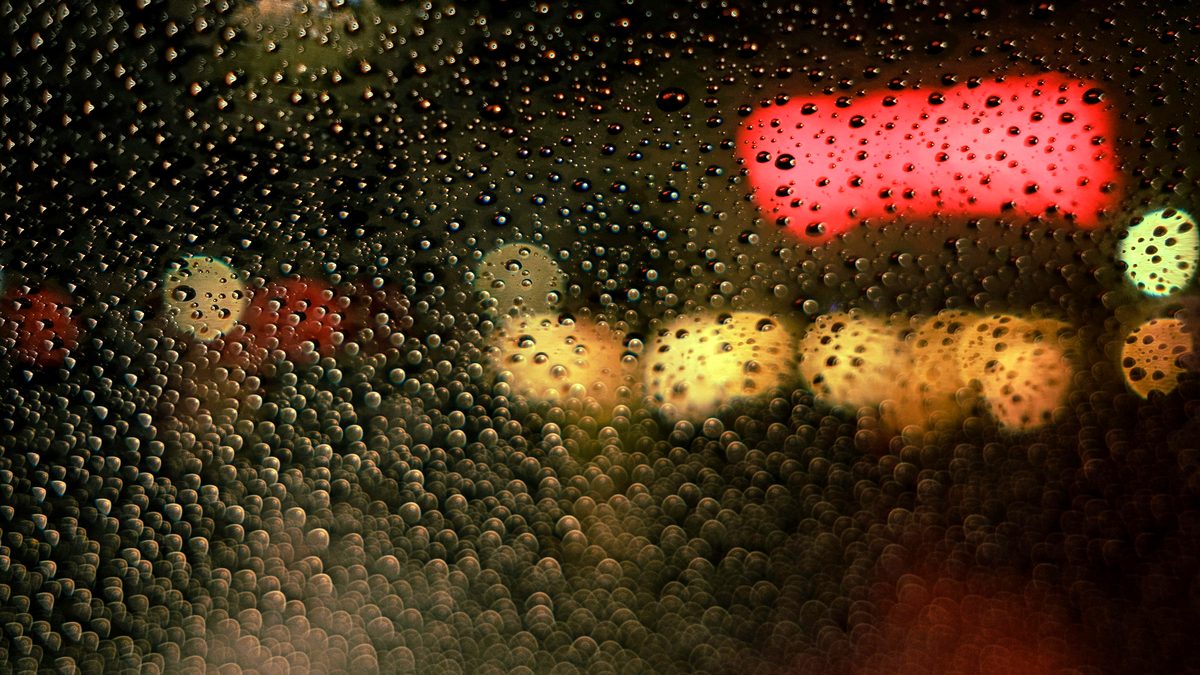 雨粒がついた窓ガラス越しに、夜の街の灯りがにじんで見えるイメージ写真